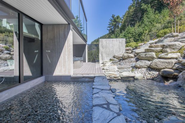 Modern architectural exterior beside a shallow landscaped water feature, with large dark-framed glazing, concrete walls, a timber deck, and a rugged natural stone stream flowing through a forested sloped site. The composition highlights the connection between the contemporary building and its rocky outdoor landscape.