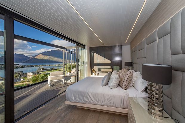 A contemporary bedroom with a large upholstered bed and padded geometric wall panels behind the headboard. The room has a white linear-panel ceiling with recessed strip lighting, dark timber-look flooring, a dark wood or charcoal feature wall with built-in joinery, and extensive black-framed sliding glazing opening onto a balcony or terrace. Through the glazing is a clear alpine lakeside landscape with mountains and waterfront buildings. Neutral grey, white and taupe furnishings create a refined, serene atmosphere.