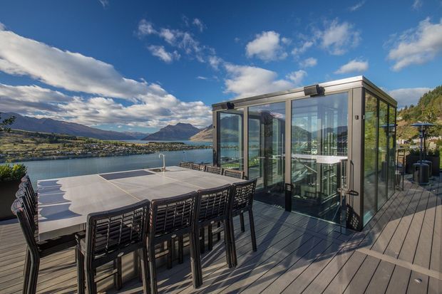 A modern glass-enclosed outdoor entertaining pavilion sits on a large timber deck overlooking a lake and mountainous landscape. The pavilion has extensive floor-to-ceiling glazing, dark metal framing, and a flat roof, while the deck includes a long dining table with bar-height chairs and an adjacent outdoor seating area. The scene is bright and scenic, with a contemporary lakeside retreat atmosphere.