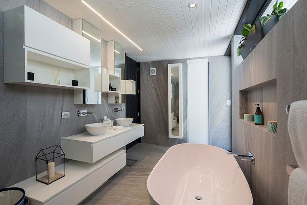 Contemporary bathroom with a freestanding pale pink bathtub in the foreground, a floating double vanity with vessel basins, mirrored and white wall cabinets, and integrated open shelving. The room features grey textured wall finishes, grey timber-look flooring, a white linear-panel ceiling, dark vertical feature cladding, recessed lighting, and indoor plants in a high-level shelf. A shower area is visible through the glazed opening at the rear.