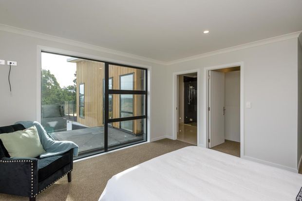 Bright contemporary bedroom with a large black-framed glazed opening overlooking a private courtyard and timber-clad structure. The room has light neutral painted walls, carpeted flooring, white bedding, and a dark upholstered armchair. A bathroom and wardrobe or storage area are visible through adjacent doorways.