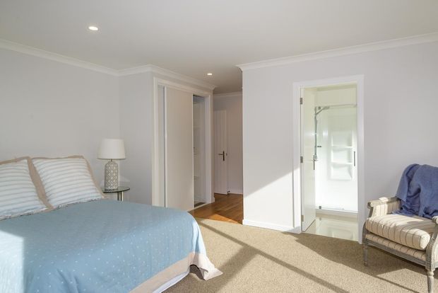 Bright, traditionally detailed bedroom with a pale blue bedspread, striped upholstered headboard and pillows, a small bedside table and lamp, and a classic upholstered armchair. White walls with decorative cornices, built-in wardrobe doors, timber flooring in the hallway, and a carpeted bedroom floor are visible. An open doorway leads to a bathroom with a glass shower enclosure and built-in shelving.