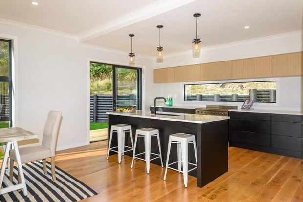 Bright contemporary open-plan kitchen and dining area with a large dark island, white bar stools, light timber flooring, extensive built-in cabinetry, pendant lighting, and an adjacent outdoor opening. A dining table and chairs are partially visible at left.