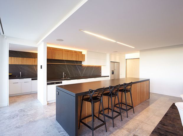 Contemporary open-plan kitchen with a large dark island and integrated seating for five, white cabinetry, warm timber overhead and island-front joinery, and a dramatic dark stone-look splashback. Linear recessed and pendant-style lighting creates a bright, refined space, with a dining or living area visible at right.