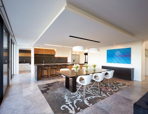 Bright open-plan contemporary interior combining a kitchen, dining area and living space. The room features a large island with timber and dark finishes, integrated white cabinetry, a long dining table with sculptural white chairs, pendant lighting, polished concrete-look flooring, and a bold blue artwork above a dark console. Full-height glazing opens along one side, creating an airy, refined atmosphere.