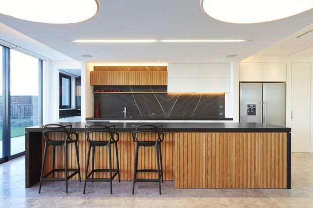 Bright contemporary kitchen with a large island featuring vertical timber-panelled cabinetry, a dark stone-look benchtop and splashback, integrated white cabinetry, black metal bar stools, and expansive glazed doors opening to an outdoor area.