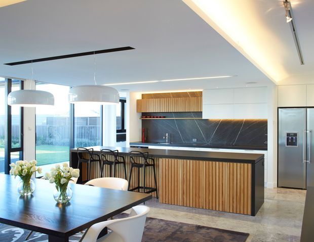 A bright, open-plan contemporary kitchen and dining area with a large timber-slat island, dark stone benchtop and splashback, integrated stainless-steel refrigerator, black-framed floor-to-ceiling glazing, pendant lights, and a dining table in the foreground. The kitchen opens to an outdoor lawn or terrace visible through the glazing.