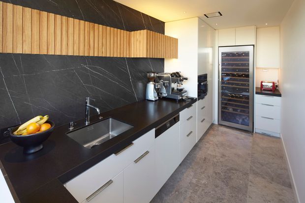 Contemporary galley kitchen with white handleless cabinetry, a dark stone-look benchtop and matching dark veined splashback, a long horizontal timber feature cabinet, stainless-steel sink and tap, integrated appliances, and a tall glass-fronted wine refrigerator. The space is compact, clean-lined, and warmly lit.