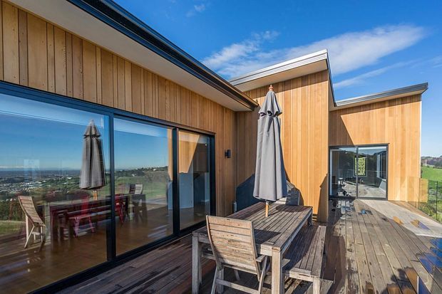 Exterior view of a modern timber-clad home with a broad timber deck and outdoor dining setting. Large dark-framed glazed doors open to the deck, while stepped roof forms and clean-lined walls create a contemporary composition. A distant landscape is visible beyond the terrace.
