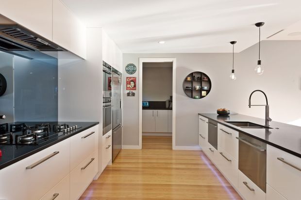 Bright contemporary galley kitchen with white flat-panel cabinetry, dark stone benchtops, a glossy blue-grey splashback, timber flooring, integrated stainless-steel appliances, pendant lighting, and a central island with a sink and curved tap. A small pantry or utility area is visible through the rear doorway.