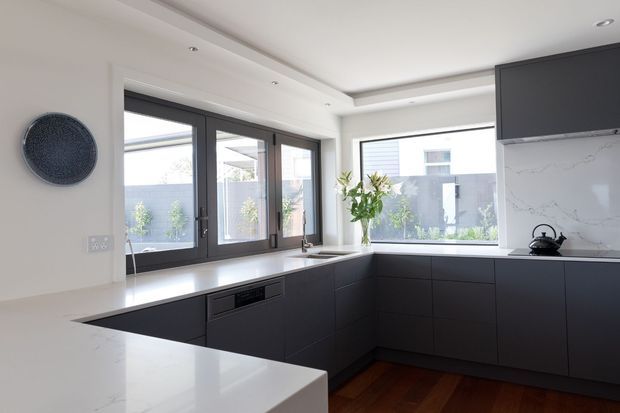 Bright contemporary kitchen with a dark matte L-shaped built-in cabinetry scheme, white stone benchtops and marble-look splashback, large dark-framed windows and doors, and warm timber flooring. The space has recessed ceiling detailing and abundant natural light.