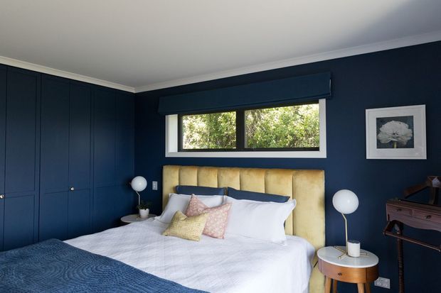 A stylish bedroom with deep navy built-in wardrobes and walls, a large bed with a pale upholstered channel-tufted headboard, white bedding, and pastel accent cushions. A horizontal window brings in greenery, while globe lamps, a framed botanical artwork, and a timber side table complete the room.