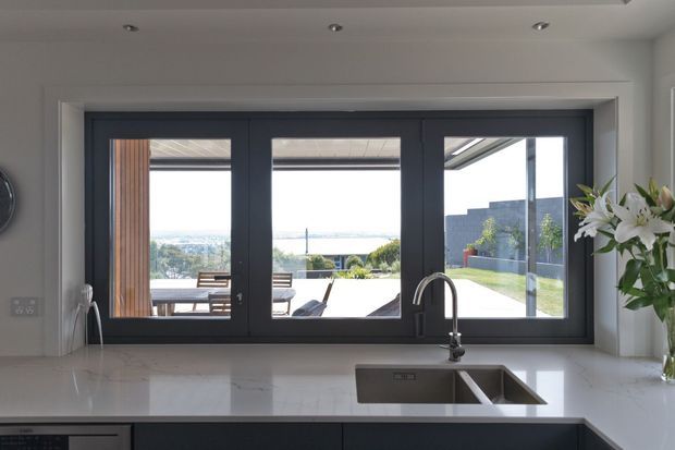 A bright, contemporary kitchen viewed from behind a white stone benchtop and deep double-bowl sink. Dark-framed folding or multi-panel glazing opens toward a spacious outdoor entertaining area with a pool or water feature and distant landscape views. The kitchen has recessed ceiling downlights, dark lower cabinetry, a minimalist tap, and a vase of lilies on the right.