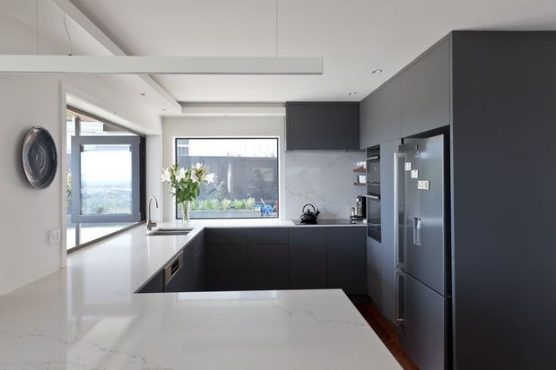 Contemporary kitchen with dark charcoal full-height cabinetry, a large white marble-look island and benchtops, integrated stainless-steel appliances, and a veined stone splashback. The bright open-plan space features generous glazing, a peninsula-style work area, and understated modern detailing.