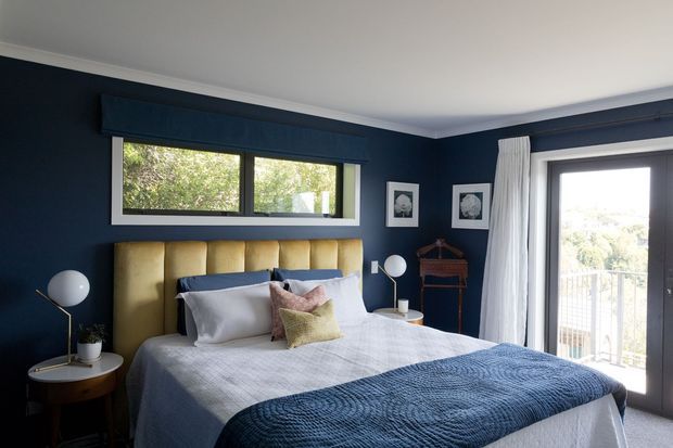 A well-appointed bedroom with deep navy walls, a large upholstered mustard-gold headboard, layered white bedding, and a textured navy throw. Dark-framed windows and a glazed balcony door bring in natural light, while white curtains, framed artwork, bedside tables, and spherical lamps add a refined contemporary character.