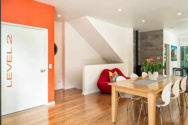 Bright contemporary dining and entry area with warm timber flooring, a light wood dining table, white sculptural chairs, a vivid red lounge chair, and a bold orange feature wall with a branded door. The room includes a geometric under-stair feature and opens toward a bright kitchen or living area.