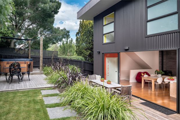Contemporary two-storey home with dark vertically ribbed cladding and large glazed openings, viewed from a landscaped backyard. A timber deck extends from the open-plan interior into the garden, with outdoor dining, a decorative outdoor cabinet, lawn, stepping stones, layered planting and dark boundary fencing. The interior visible through the opening includes timber flooring, a dining table and chairs, and a bright sitting area with a red lounge chair.