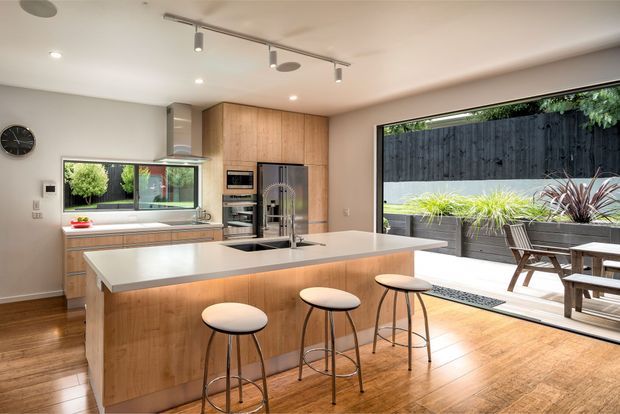 Bright contemporary kitchen with extensive timber cabinetry, a large white island with integrated sink and seating for four, stainless-steel appliances, timber flooring, and expansive glazing opening to a landscaped outdoor dining area.