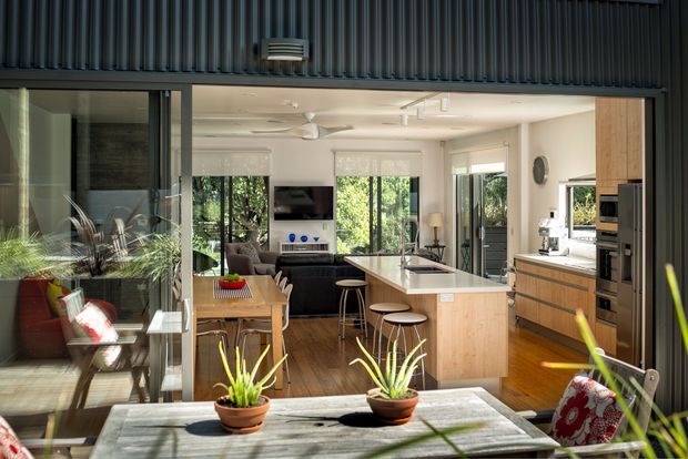 A contemporary open-plan home interior viewed through a glazed living area, with a bright kitchen featuring a large island, timber cabinetry, integrated appliances and pendant lighting. The adjoining living and dining spaces have timber floors, dark seating, indoor plants and extensive glazing. A dark corrugated exterior or ceiling-like cladding element frames the upper portion of the image.