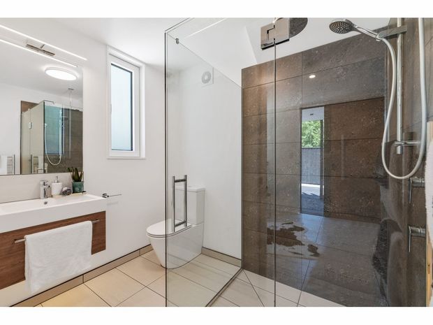Bright contemporary bathroom with a frameless glass shower enclosure, large dark stone-look wall tiles, pale floor tiles, a wall-mounted timber vanity with white basin, mirrored cabinetry, and a wall-hung toilet. Natural light enters through a narrow window, creating a clean, refined space.