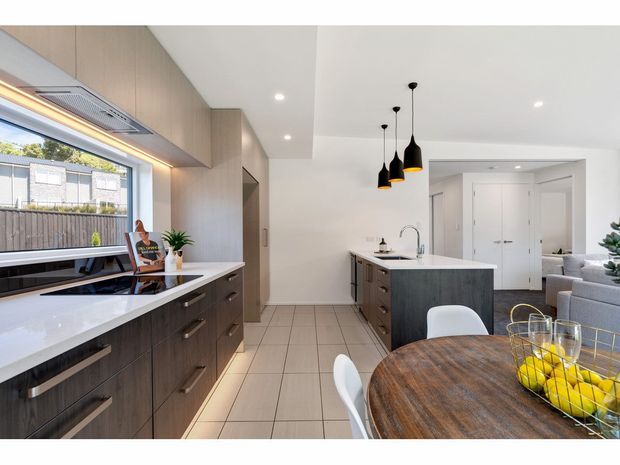 Bright contemporary open-plan kitchen and dining area with dark timber-look cabinetry, white stone benchtops, a central island, integrated appliances, black pendant lights and a round timber dining table. Large glazing brings in natural light and overlooks a neighbouring residential setting.