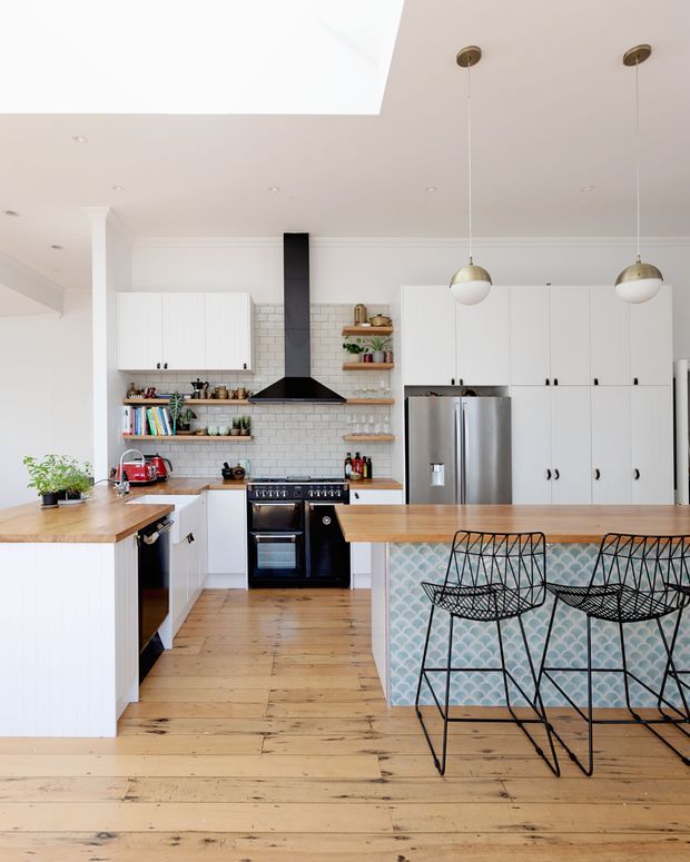 Bright, spacious white kitchen with timber flooring and timber benchtops, extensive white cabinetry, a stainless-steel refrigerator and freestanding black range cooker. A white subway-tile splashback is paired with open timber shelving, while the island features a pale blue scallop-patterned front and black wire bar stools. Brass and opal pendant lights hang from a high white ceiling.