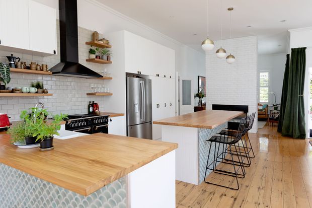 Bright, open-plan kitchen with white cabinetry, subway-tile splashback, black rangehood and appliances, timber benchtops, and a patterned scallop tile feature on the island. A second island or breakfast bar with black wire stools separates the kitchen from the adjoining living area. Pendant lights, timber floorboards, indoor plants, and dark green curtains add warmth and contrast.