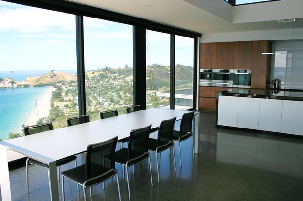 A contemporary open-plan kitchen and dining area with a long white dining table and black chairs positioned beside full-height black-framed glazing. The kitchen features white island cabinetry, dark stone-like benchtops, timber veneer wall cabinetry, integrated stainless-steel ovens, and a polished grey floor. The glazing overlooks a bright coastal bay and surrounding hills.