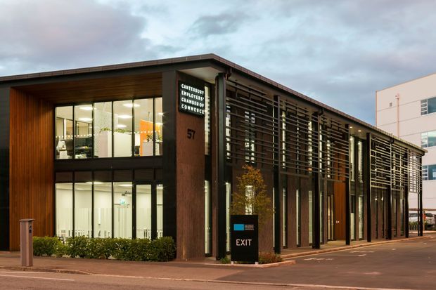 Exterior view of a contemporary two-storey commercial building with a long, low flat roof, dark timber-toned cladding, extensive glazing, and a prominent horizontal timber or metal screen along the facade. The entrance is marked with signage and an exit sign, while a lighter neighbouring commercial building is visible at right. The scene is photographed at dusk under an overcast sky.