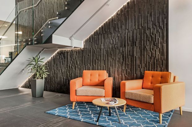 Contemporary reception or lounge area beneath a glass-and-metal staircase, featuring two orange upholstered armchairs, a small round timber coffee table, and a blue patterned rug. A dramatic textured dark feature wall with integrated linear lighting forms the backdrop, while a potted plant adds greenery.