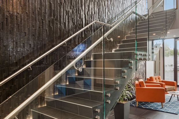 A contemporary interior stairway with dark tiled treads and risers, glass balustrades, stainless-steel handrails, and a dramatic textured dark feature wall. An orange lounge seating area and large windows are visible beyond the stairs.