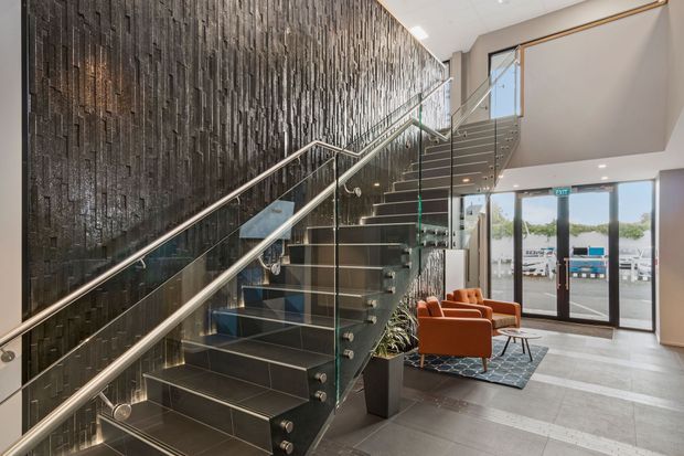 A contemporary commercial-style entry lobby dominated by a wide staircase with dark tiled or stone-clad treads, stainless-steel handrails, and clear glass balustrades. A dramatic textured dark feature wall runs alongside the stairs, while a small seating area with rust-coloured lounge chairs sits near full-height glazed entry doors. The space is bright, polished, and highly refined.