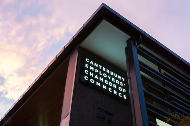 Low-angle exterior view of a contemporary commercial building at dusk, featuring a sharply pitched roofline, dark timber-like wall cladding, exposed structural framing, horizontal balcony or facade rails, and an illuminated sign reading “Canterbury Employers’ Chamber of Commerce.”