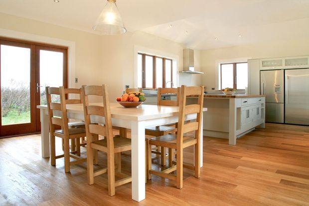 Bright open-plan kitchen and dining area with a large white island, integrated seating, timber dining table and chairs, stainless-steel appliances, cream cabinetry, timber-framed glazing, and warm timber flooring. The room has abundant natural light and a clean, functional family-oriented layout.