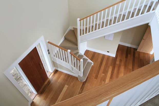 Elevated view of a bright residential stairway and landing with white-painted balustrades, warm timber handrails, stained timber flooring, and carpeted stairs. A timber entry door and hallway are visible below.
