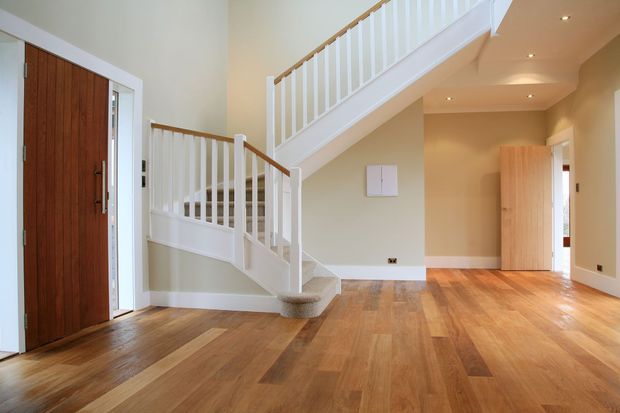 Bright residential entry hall with warm timber flooring, a substantial timber front door, and a white-painted staircase with turned newel posts and balustrades. Beige walls, white skirtings, recessed ceiling lights, and an open doorway create a clean, welcoming transitional space.