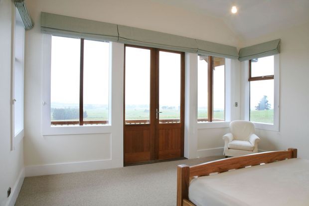 Bright traditional bedroom with a timber-framed glazed door and large windows overlooking open countryside. The room has pale painted walls with simple panel moulding, carpeted flooring, muted green Roman blinds, and a timber bed with a cream upholstered armchair. The space is airy and lightly furnished.