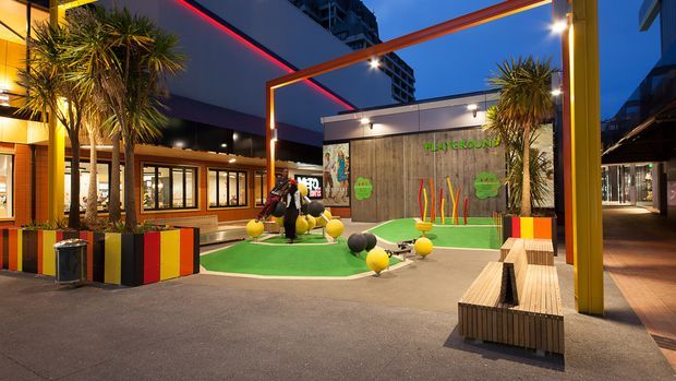A vibrant urban outdoor recreation courtyard at dusk, featuring a colourful mini-golf putting area with artificial turf, playful oversized golf-ball ornaments, striped planter boxes, palm-like plants, timber seating, and a mural-style feature wall. Contemporary buildings and illuminated architectural elements frame the space, creating a lively hospitality or commercial entertainment setting.