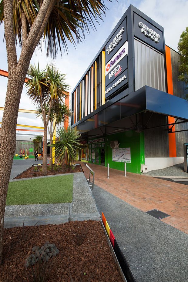 Exterior view of a contemporary commercial complex with a prominent dark charcoal and orange facade, projecting blue canopy, vertical fins, and multiple business signs. The building fronts a landscaped pedestrian approach with palm-like plants, raised garden beds, synthetic turf, brick paving, and accessible-style handrails.
