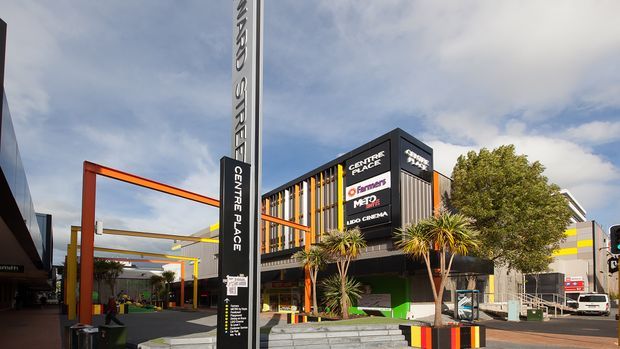 Exterior view of a contemporary multi-level commercial shopping and entertainment complex, with prominent Centre Place signage, colourful orange and yellow structural frames, dark cladding, retail and cinema tenant signs, landscaped entry planting, and a paved pedestrian forecourt under a partly cloudy sky.