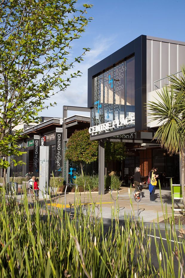 Exterior streetscape view of a contemporary commercial dining precinct, with dark framed retail and restaurant buildings, large glazed façades featuring decorative line graphics, vertical signage, landscaped planting, and pedestrian walkways. The prominent entrance signage identifies a dining venue, while adjacent storefronts and outdoor seating create an active hospitality setting.