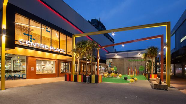 An urban commercial shopping and entertainment plaza at dusk, featuring the illuminated Centre Place storefront, bold contemporary facade signage, colourful geometric planter boxes, sculptural palm plantings, and a framed outdoor public space with miniature-golf-style landscaping and seating. The scene is brightly lit with vivid yellow, red, orange, and green accents against a deep blue evening sky.