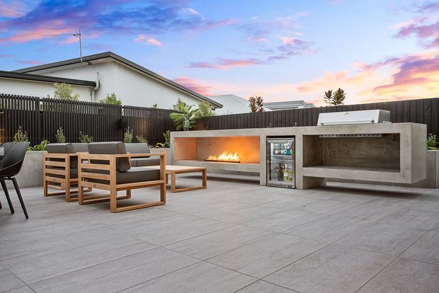 Contemporary outdoor entertaining area with a large tiled patio, timber outdoor lounge seating, a built-in concrete fireplace, integrated outdoor kitchen or bar cabinetry, and a compact beverage refrigerator. A dark vertical fence encloses the space, with neighbouring pitched-roof homes visible beyond it at sunset.