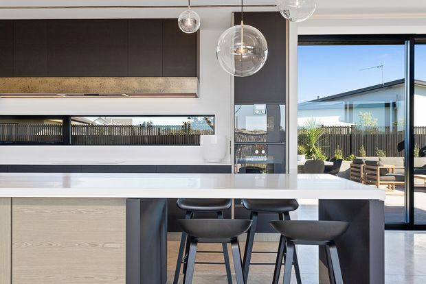 Contemporary open-plan kitchen with a large white island and integrated seating, light timber cabinetry, dark upper cabinetry, a stone or engineered-stone benchtop, and spherical glass pendant lights. Full-height glazing opens to an outdoor terrace and shows a neighbouring modern home and fence beyond.