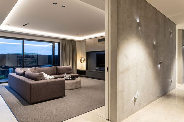 A refined contemporary living room viewed from behind a textured feature wall. The room contains a large taupe sectional sofa, round upholstered ottoman coffee table, integrated dark entertainment cabinetry, recessed ceiling lighting and full-height glazing opening to an outdoor terrace. The feature wall has a raw concrete-like finish with small directional wall lights, while light stone or porcelain flooring continues through the circulation area. The space is softly lit, neutral-toned and highly finished.