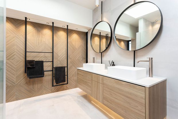 Contemporary double-vanity bathroom with a walk-in shower, featuring light timber-look cabinetry, white vessel basins, large round black-framed mirrors, pale wall tiling, and a geometric timber-toned feature wall with black heated towel rails.