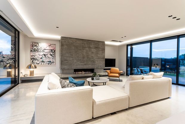 A spacious contemporary living room with polished concrete flooring, white modular sofas, a dark stone feature wall with a linear fireplace and television, and extensive floor-to-ceiling black-framed glazing opening to an outdoor terrace. Recessed lighting, sculptural artwork, a low coffee table, and warm leather seating create a refined minimalist interior with strong indoor-outdoor connection.