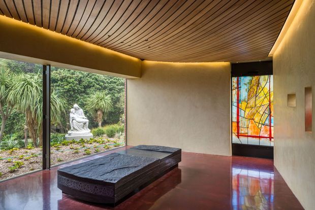A refined interior gallery or entry space with a large dark stone-topped display plinth or table, polished reddish-brown flooring, curved warm plaster walls, and a dramatic curved timber-slat ceiling with concealed lighting. Full-height glazing opens views to a landscaped garden and sculpture, while a colourful abstract stained-glass or leadlight panel forms a feature beside the doorway. The space has a calm, gallery-like atmosphere with strong natural and warm accent lighting.