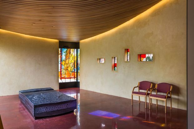 A quiet, gallery-like interior with curved plaster walls, a richly stained polished floor, a ribbed timber ceiling, and concealed warm cove lighting. A large sculptural dark upholstered bench or ottoman sits in the foreground, while two burgundy chairs line the wall. A tall stained-glass door or window and several small stained-glass wall insets introduce vivid reds, yellows, blues, and greens.