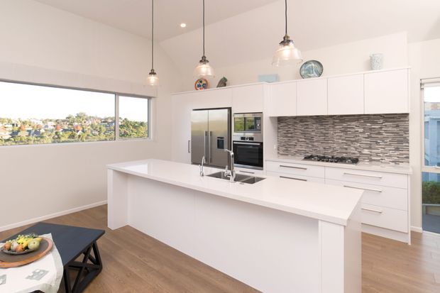 Bright contemporary white kitchen with a large island and integrated sink, white flat-panel cabinetry, stainless-steel appliances, and a grey stacked mosaic tile splashback. Three glass pendant lights hang above the island, while timber flooring and expansive windows add warmth and natural light. A dining area is partially visible in the foreground.